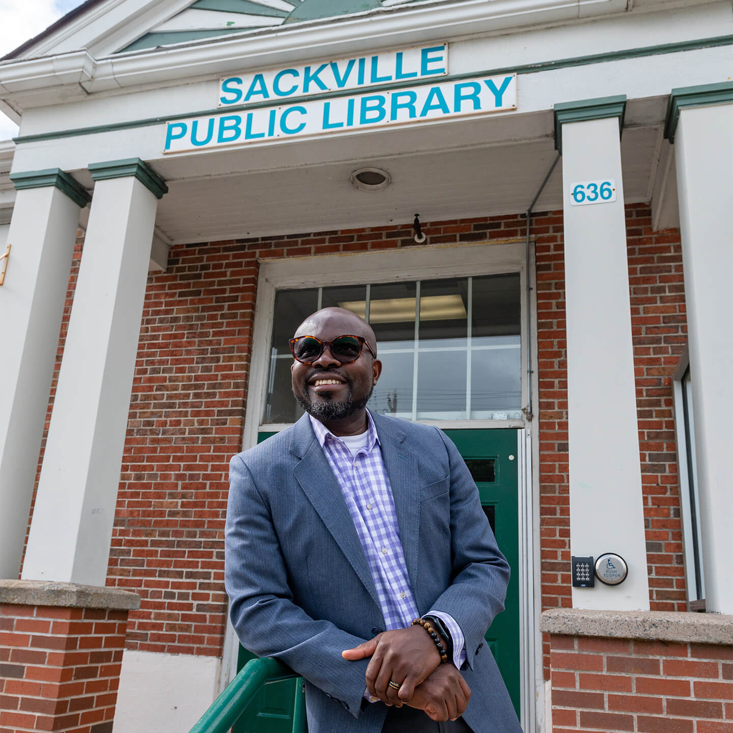 Ayo is wearing a blue suit and sunglasses, leaning on the railing in front of the Sackville Public Library entrance.